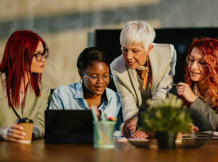 Senior businesswoman is standing at boardroom with her multicultural team and tutoring them. Mature female mentor is teaching her interracial team new business strategies and analyzing data with them.