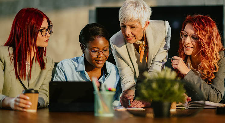 Senior businesswoman is standing at boardroom with her multicultural team and tutoring them. Mature female mentor is teaching her interracial team new business strategies and analyzing data with them.