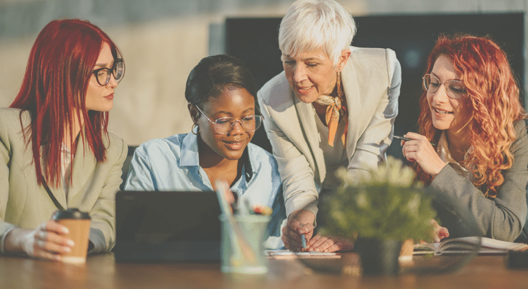 Senior businesswoman is standing at boardroom with her multicultural team and tutoring them. Mature female mentor is teaching her interracial team new business strategies and analyzing data with them.